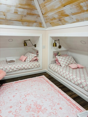 A room with two bunk beds, each with a bedside lamp, a small table, and a pink floral rug.