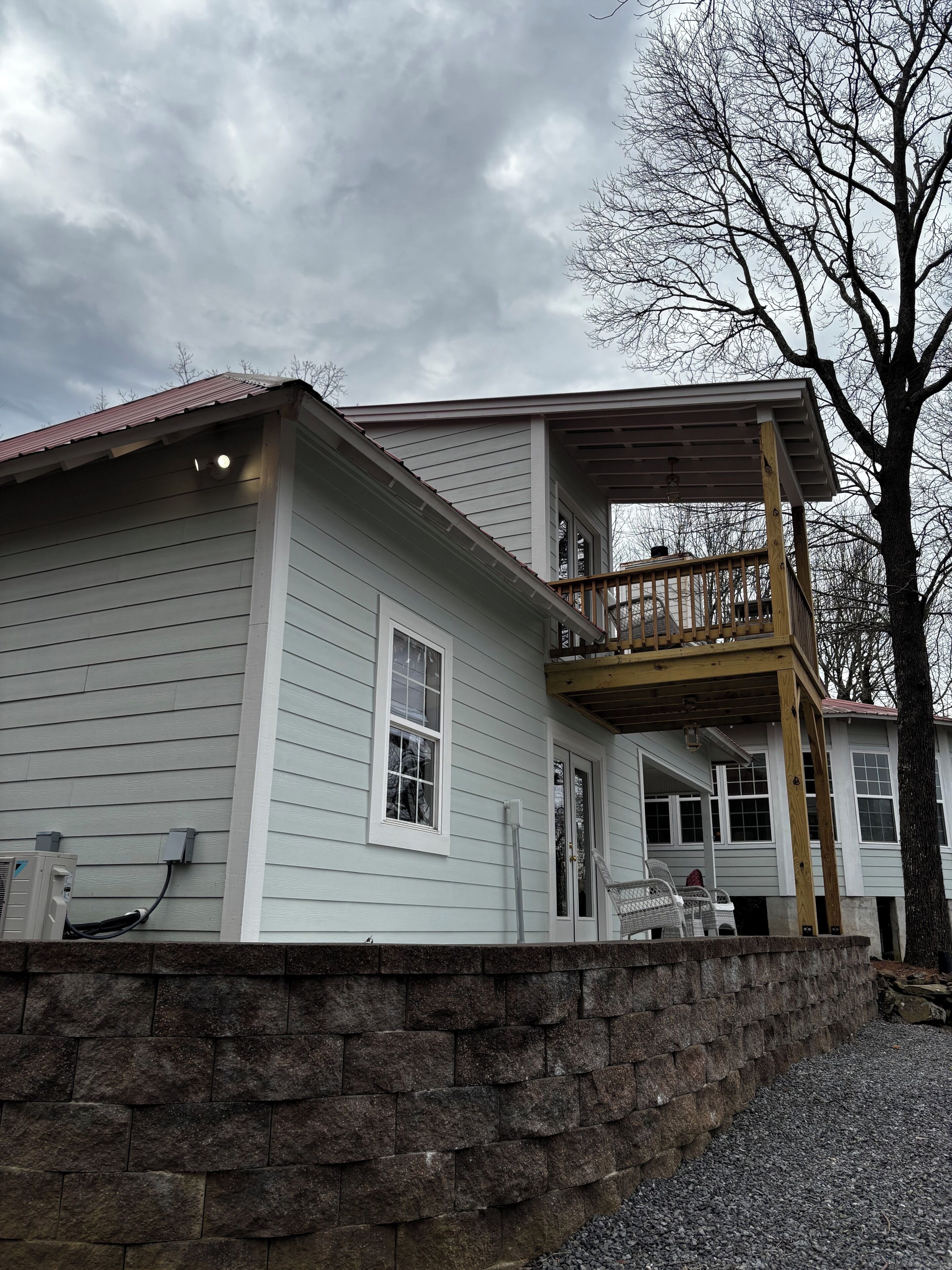 A two-story house with a balcony, a stone retaining wall, and a cloudy sky.