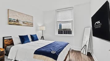 A hotel room with a bed, a desk, a lamp, a window, and a painting on the wall.