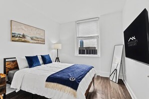 A hotel room with a bed, a desk, a lamp, a window, and a painting on the wall.