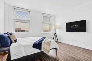 Habitación de hotel con cama, dos ventanas, una lámpara de pie y un televisor empotrado en la pared que muestra 'TRAVELLER'S COMFORT'.