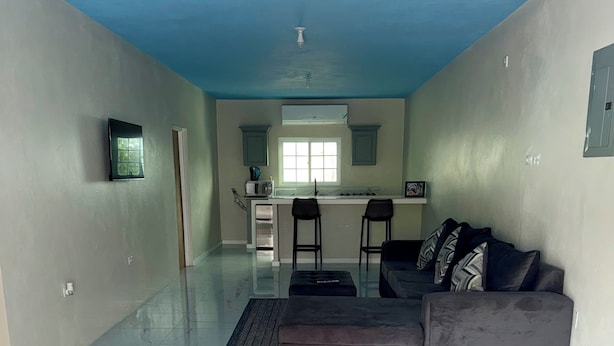 Amplia sala de estar con un sofá y reposapiés, una cocina con ventana y suelo de baldosas.