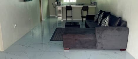 A spacious living area with a sofa and ottoman, a kitchen with a window, and a tiled floor.