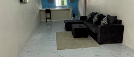 A living room with a sofa, a coffee table, and a TV mounted on the wall. There is a kitchen area in the background with a window and a bar stool.