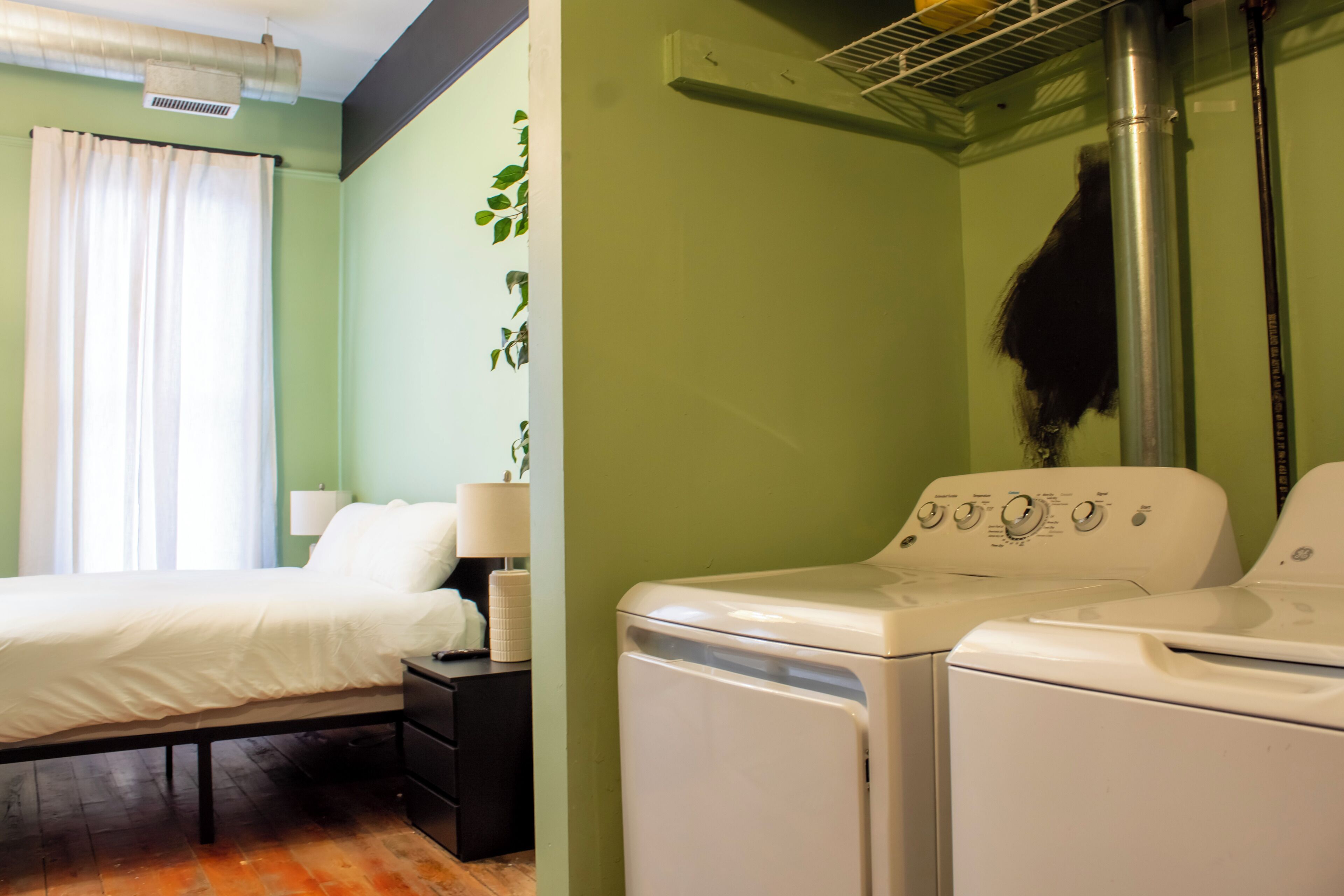 A room with a bed, a nightstand, a lamp, a wall-mounted heater, and a washing machine.