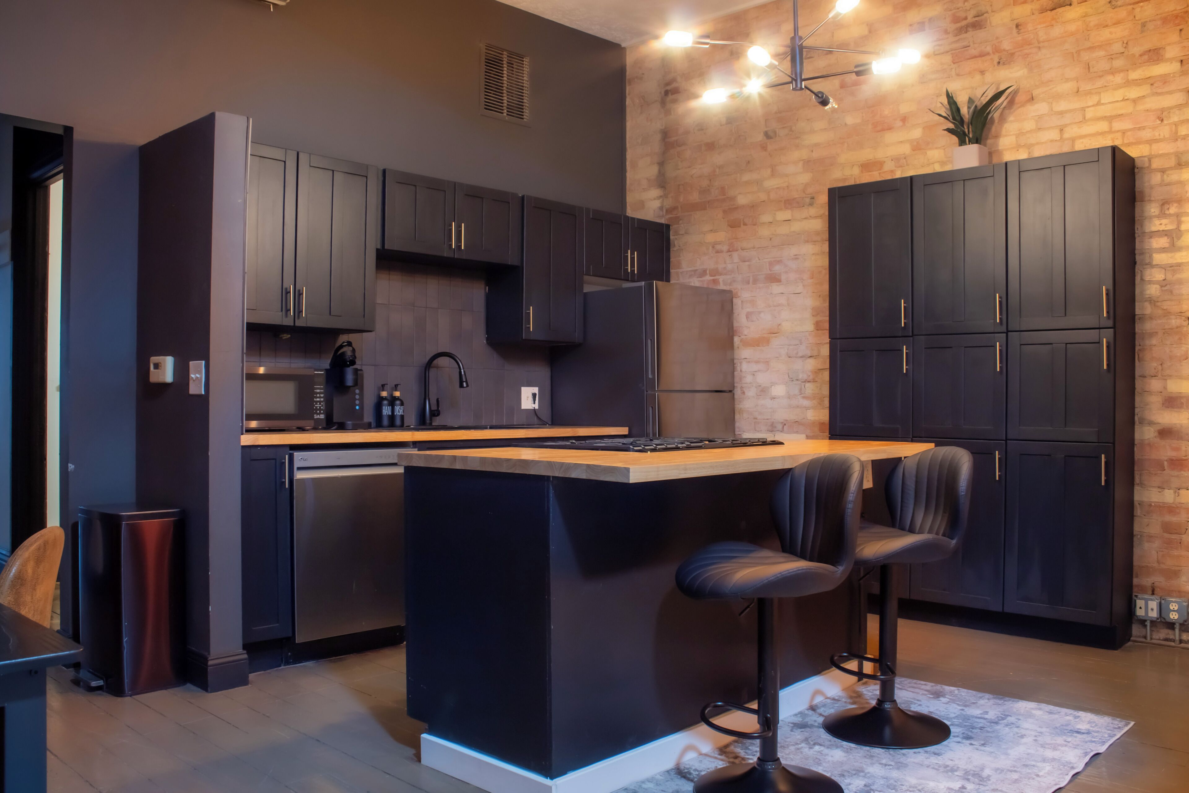 A modern kitchen with dark cabinetry, a central island with bar stools, and a brick wall.