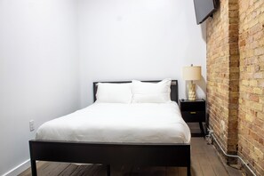 Una cama individual con ropa blanca en una habitación con pared de ladrillo y un televisor de pantalla plana montado encima.