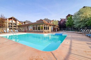 A swimming pool with lounge chairs, a building with a covered patio, and a fenced area with trees. - The Current | Outside Pool | Hot Tub | Gym | Pets (Invermere)