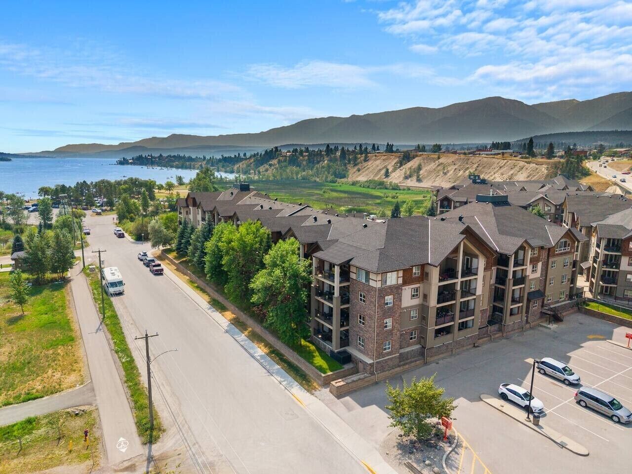 A multi-story building with a parking lot, surrounded by greenery and a view of a lake and mountains.