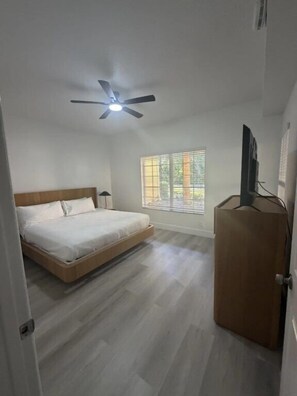 Een slaapkamer met een bed, een plafondventilator, een raam met jaloezieën en een houten deur.