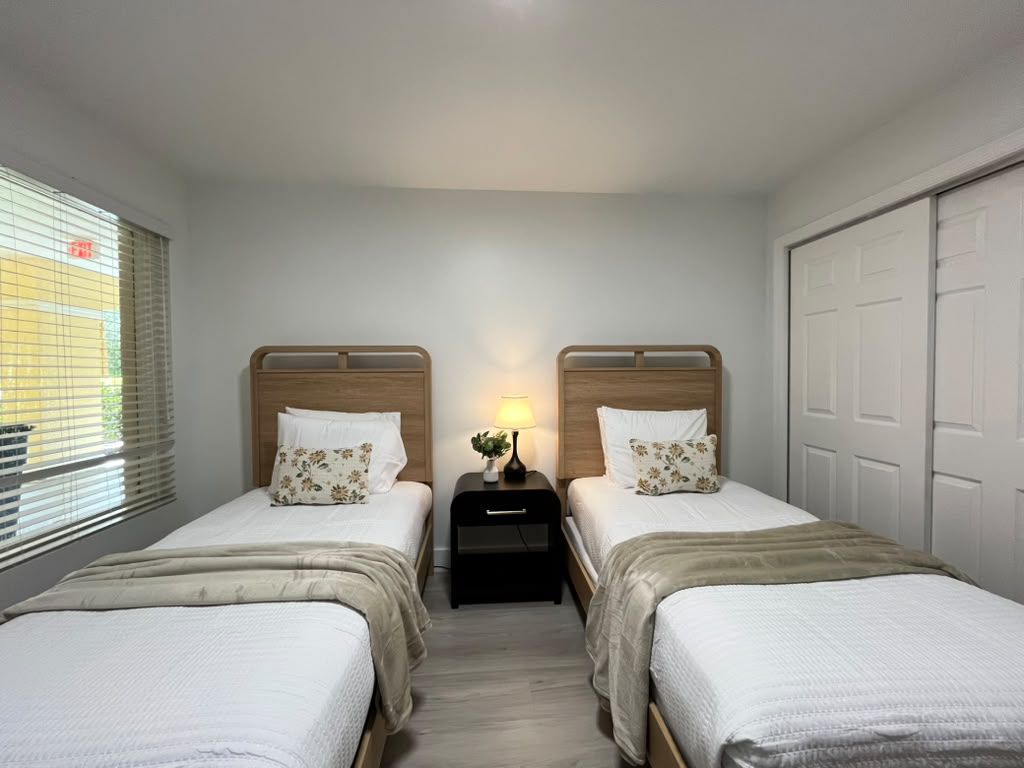 A room with two beds, a nightstand with a lamp, and a window with blinds.