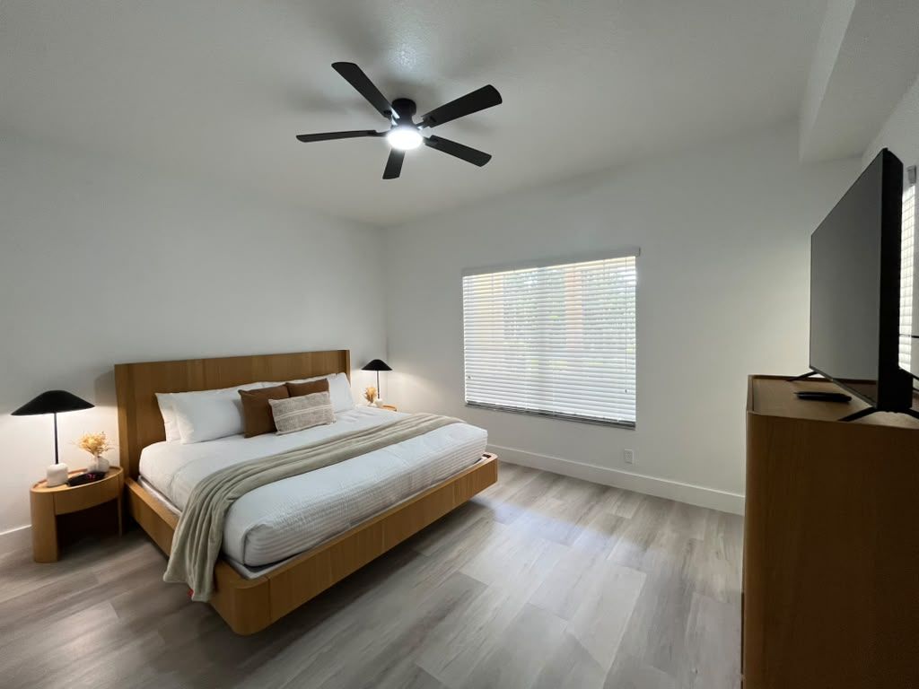 Ein Schlafzimmer mit Bett, Nachttischen, Deckenventilator, Fenster mit Jalousien und einem Fernsehmöbel.