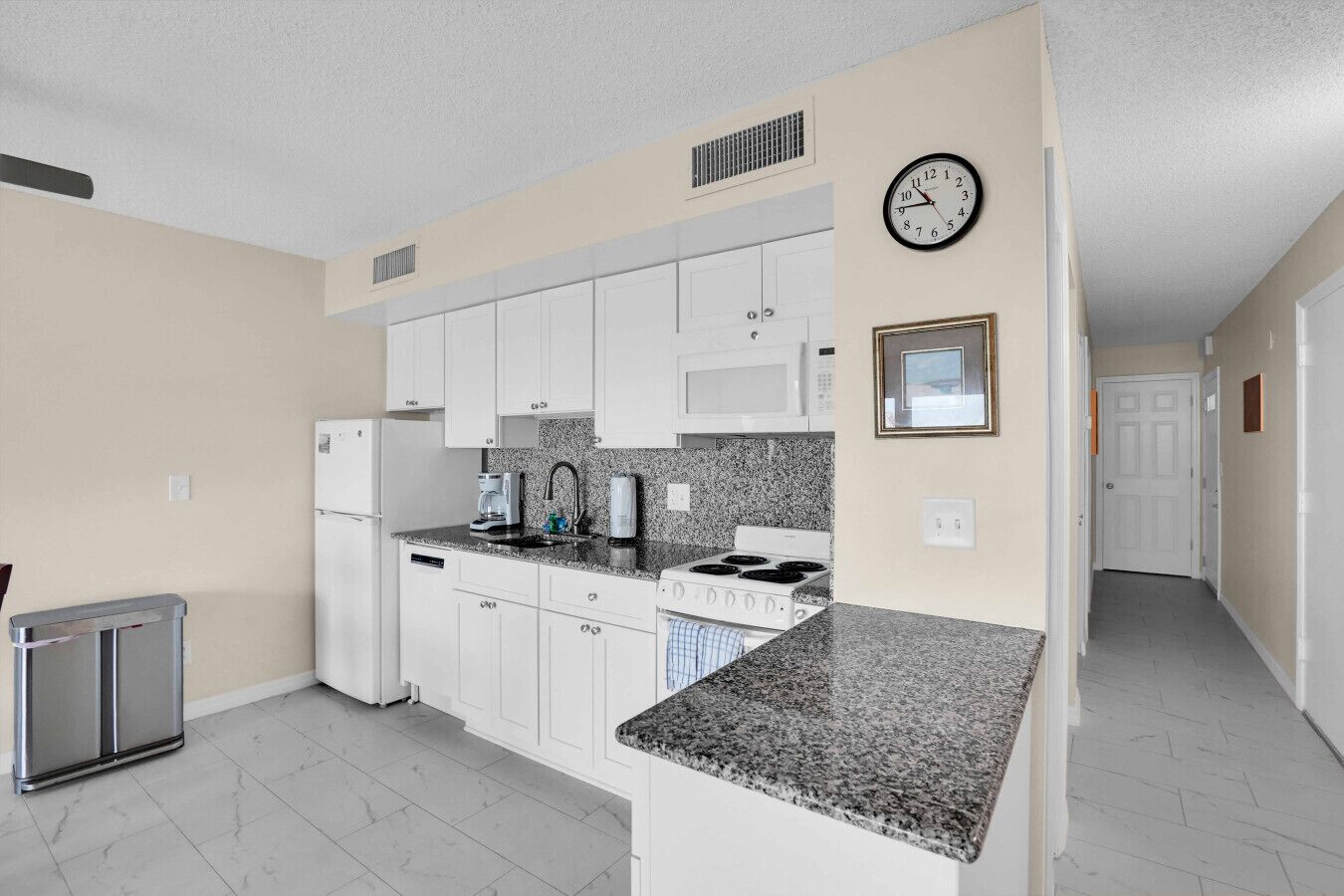 A kitchen with white cabinets, a granite countertop, a microwave, a stove, and a clock on the wall.