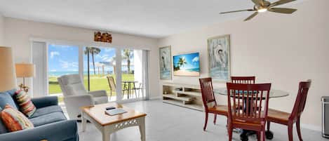A spacious living room with a sofa, coffee table, and dining area with chairs. There is a ceiling fan and artwork on the walls.