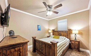 Ein Schlafzimmer mit einem Bett, Holz-Nachttischen, einem Deckenventilator, einem Fernseher und einem Wandventilator.