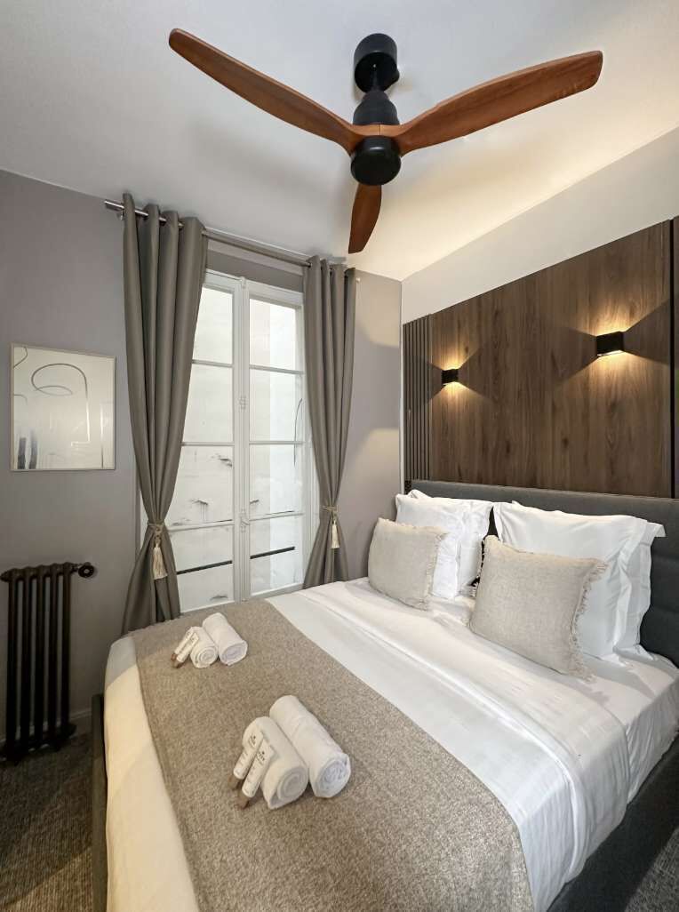 Une chambre d’hôtel avec un lit, un ventilateur de plafond, une fenêtre avec des rideaux et un luminaire fixé au mur.