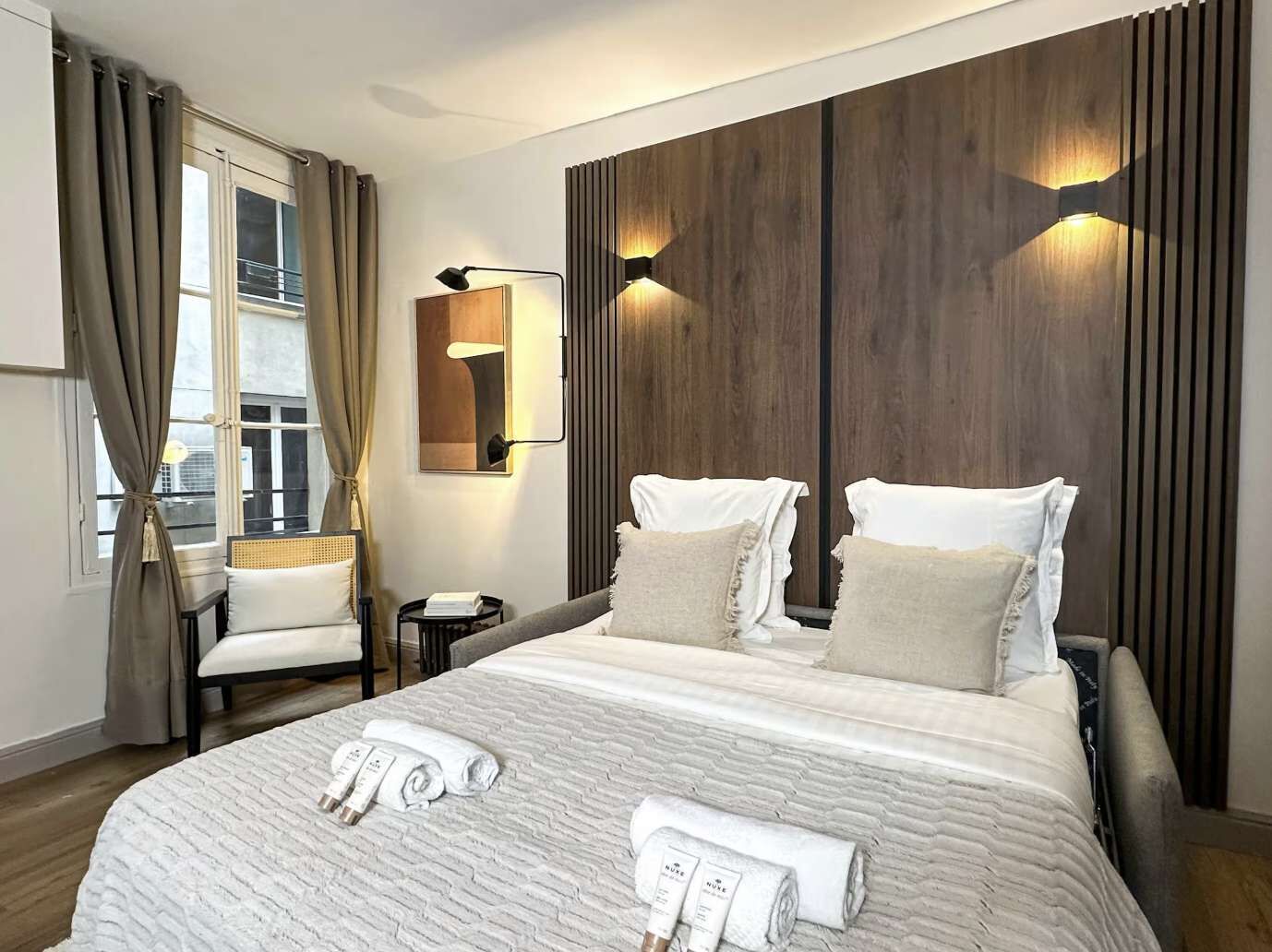 A hotel room with a large bed, a bedside table, a chair, a window with curtains, and a wall-mounted light fixture.