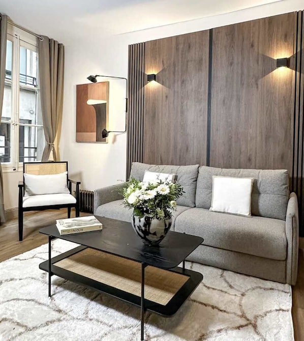 Un salon moderne comprenant un canapé, une table basse, un fauteuil, un vase de fleurs et un luminaire fixé au mur.