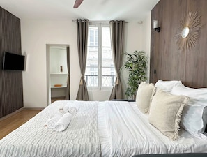 Ein Hotelzimmer mit Bett, Fernseher, Fenster mit Vorhängen, einer Pflanze und einem Wandspiegel.