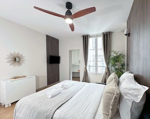 Ein Schlafzimmer mit Bett, Deckenventilator, Fernseher, einem Fenster mit Vorhängen, einer Pflanze und einem Heizkörper.