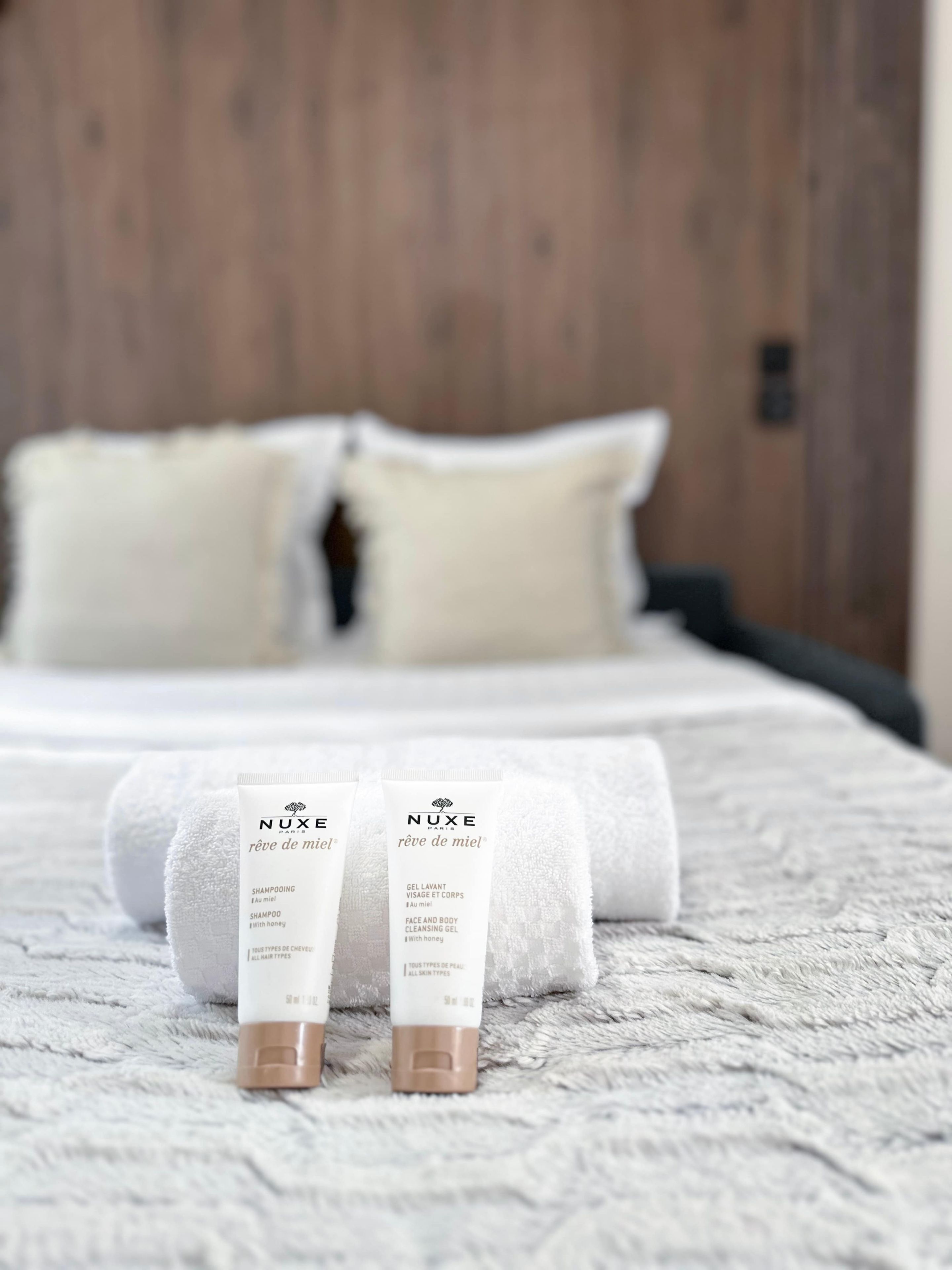 A neatly made bed with white linens and two Nuxe skincare products placed on the bed.