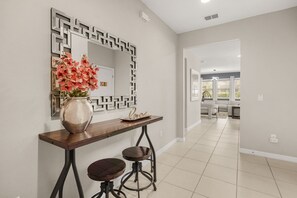 Een modern interieur met een eethoek bestaande uit een houten tafel, twee krukken en een vaas met bloemen. Aan de muur hangt een decoratieve spiegel met een geometrisch frame.