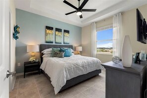 A bedroom with a bed, bedside lamps, a ceiling fan, artwork on the wall, a window with a view, and a TV mounted on the wall. - 2 BD/ 2 BA/ Storey Lake (3141-404 PC) (Kissimmee)