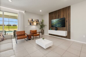 A modern living room with a flat-screen TV mounted on a wooden panel wall, two leather armchairs, a white coffee table, and a large window overlooking a green landscape. - 10 BD/ 8 BA/ Windsor Island Resort (4321 LA) (Davenport)