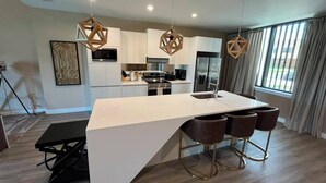 A modern kitchen with a central island, bar stools, pendant lighting, and a large window with curtains. - 3 BD/ 3.5 BA/ Magic Village by Pininfarina (5591 PW) (Kissimmee)