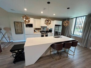 Een moderne keuken met een centraal eiland, barstoelen, hanglampen en een groot raam met gordijnen.