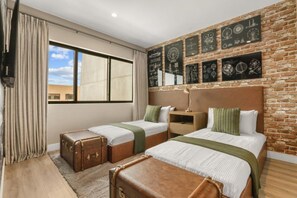 Ein Hotelzimmer mit zwei Betten, einer Ziegelwand mit gerahmten Bildern, einem Fenster mit Vorhängen und einem Koffer auf dem Boden.