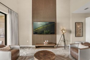 A modern living room with a large flat-screen TV mounted on a wooden panel wall, leather sofas, a coffee table, and a tripod lamp. - Brand New! 3 Bd/3.5 Ba/ Magic Village by Pininfarina - 5591 PW (Kissimmee)