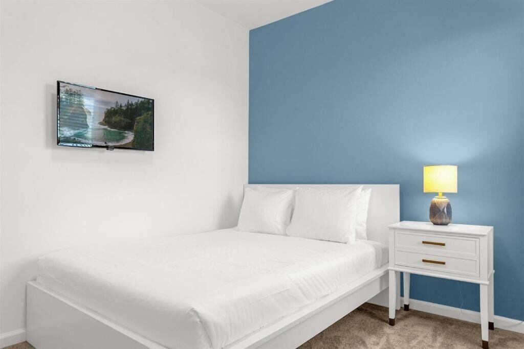 A bedroom with a bed, a nightstand with a lamp, and a wall-mounted TV displaying a landscape.