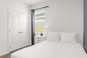 Ein Schlafzimmer mit einem Bett, einem Nachttisch mit Lampe, einem Fenster mit Jalousien und einer Tür.