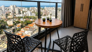 A balcony with a round table and two chairs, overlooking a cityscape with buildings and a clear sky. - Pause Living - Beach Class Rio Vermelho (Salvador)