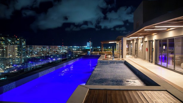A rooftop pool with a city view, a covered seating area, and a modern building facade. - Pause Living - Beach Class Rio Vermelho (Salvador)