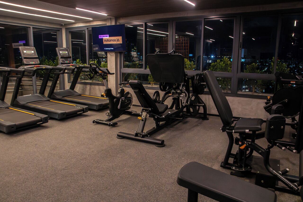 A gym with treadmills, stationary bikes, and weight machines, with a city view through large windows.