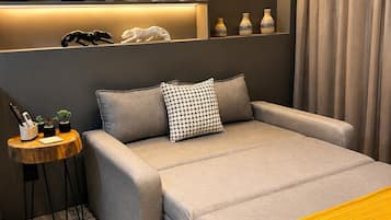 A modern bedroom with a sofa bed, a small side table, a yellow throw blanket, and a wall-mounted shelf with decorative items.