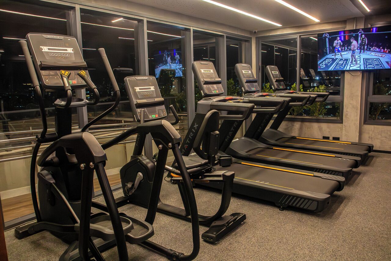 Une salle de sport équipée de tapis de course et d’appareils d’exercice, dotée de grandes fenêtres et d’un écran de télévision diffusant un événement sportif.