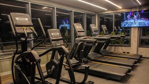 A gym with treadmills and exercise equipment, large windows, and a TV screen displaying a sports event. - Pause Living - Beach Class Rio Vermelho (Salvador)