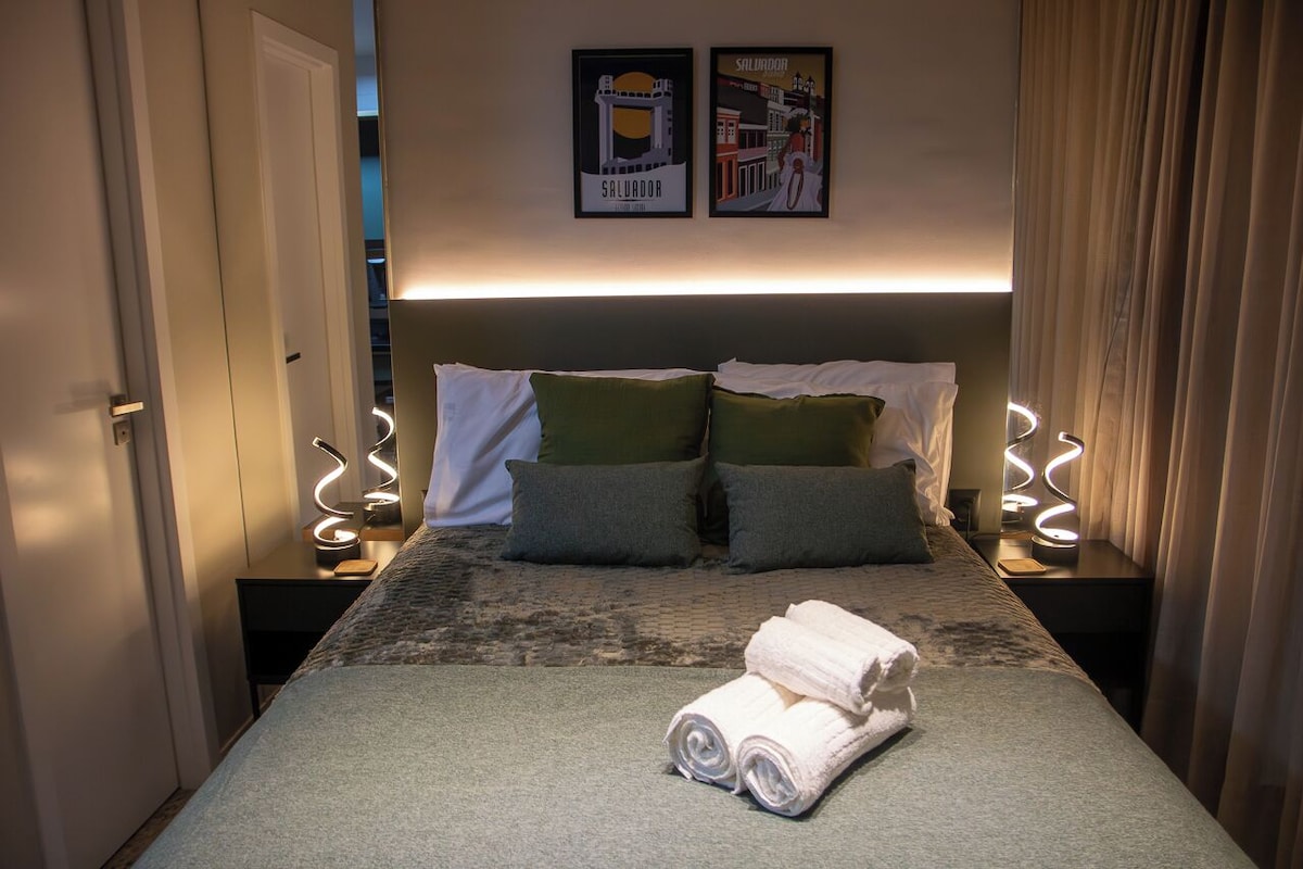 Een hotelkamer met een bed, twee kussens, een keurig opgevouwen handdoek en twee wandlampen.