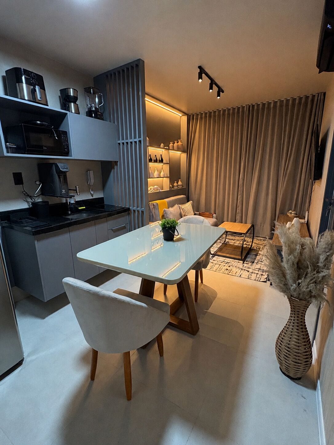 Una cocina moderna con electrodomésticos integrados, un comedor con mesa y sillas blancas, y una sala de estar con sofá y cortinas.