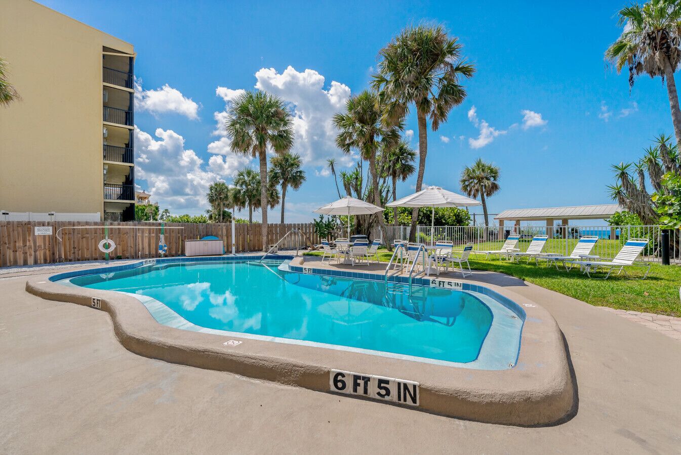 A swimming pool with lounge chairs and umbrellas, surrounded by palm trees and a building.