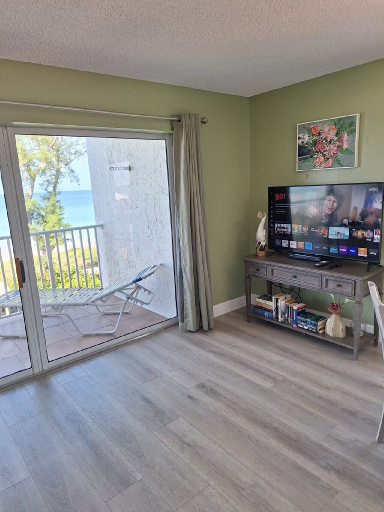 Een kamer met een schuifglasdeur die uitkomt op een balkon, een televisie op een meubelstuk en een kleine tafel met boeken en decoratieve objecten.