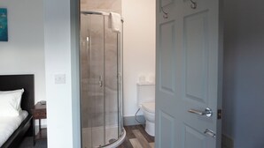 Hotellihuone, jossa on sänky, yöpöytä, lasiovilla varustettu suihku, wc ja ovi.