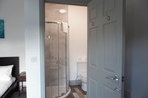 Quarto de hotel com cama, mesa de cabeceira, chuveiro com porta de vidro, vaso sanitário e porta.