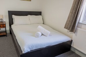 Cama bem arrumada com lençóis brancos e uma toalha enrolada, cabeceira escura e uma janela com cortina.