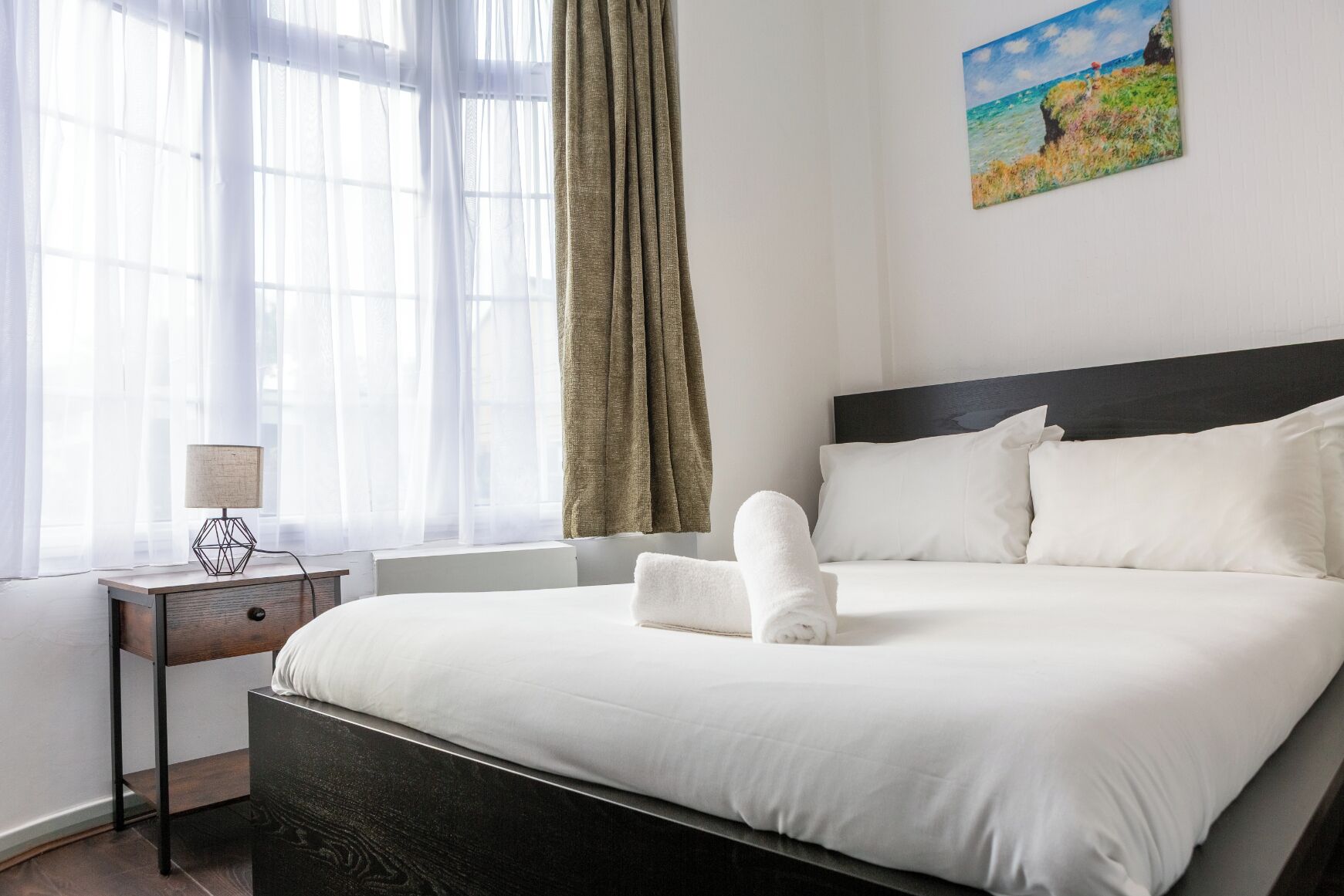 A hotel room with a large bed, a bedside table with a lamp, a window with sheer curtains, and a painting on the wall.