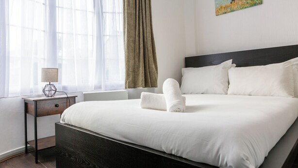 Hotellihuone, jossa on suuri sänky, yöpöytä lampun kanssa, ikkuna, jossa on läpikuultavat verhot, ja taulu seinällä.