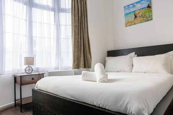 Quarto de hotel com uma cama grande, mesa de cabeceira com abajur, janela com cortinas finas e um quadro na parede.